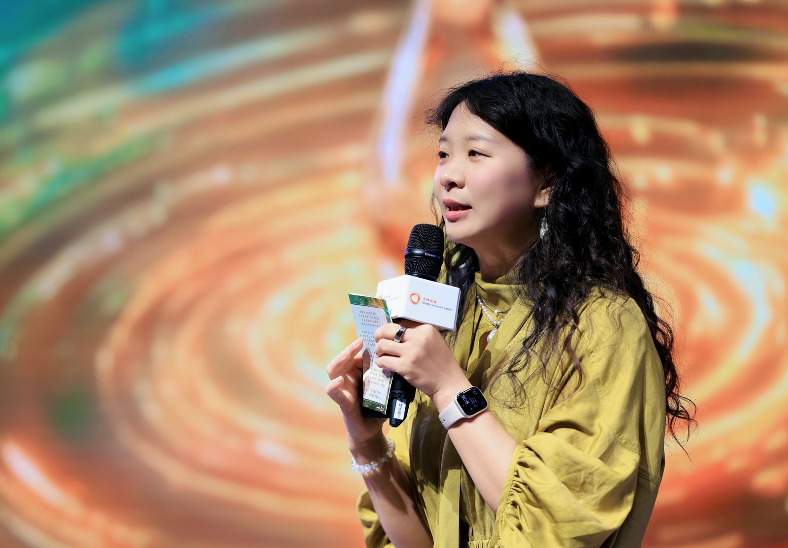 Marcia speaking warmly, microphone in hand, against golden ripples