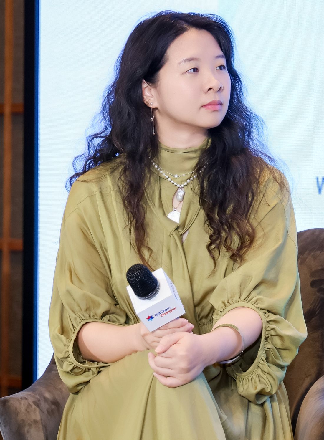 Marcia seated on stage in an olive dress, listening